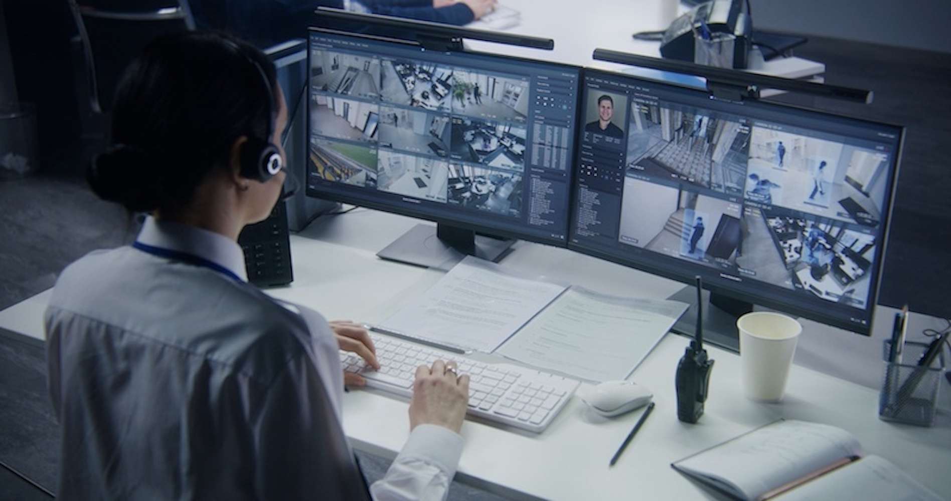 Person wearing a headset monitors multiple security camera feeds on two large computer screens at a desk with documents, a keyboard, a walkie-talkie, a coffee cup, and a notebook. Person wearing a headset monitors multiple security camera feeds on two large computer screens at a desk with documents, a keyboard, a walkie-talkie, a coffee cup, and a notebook.
