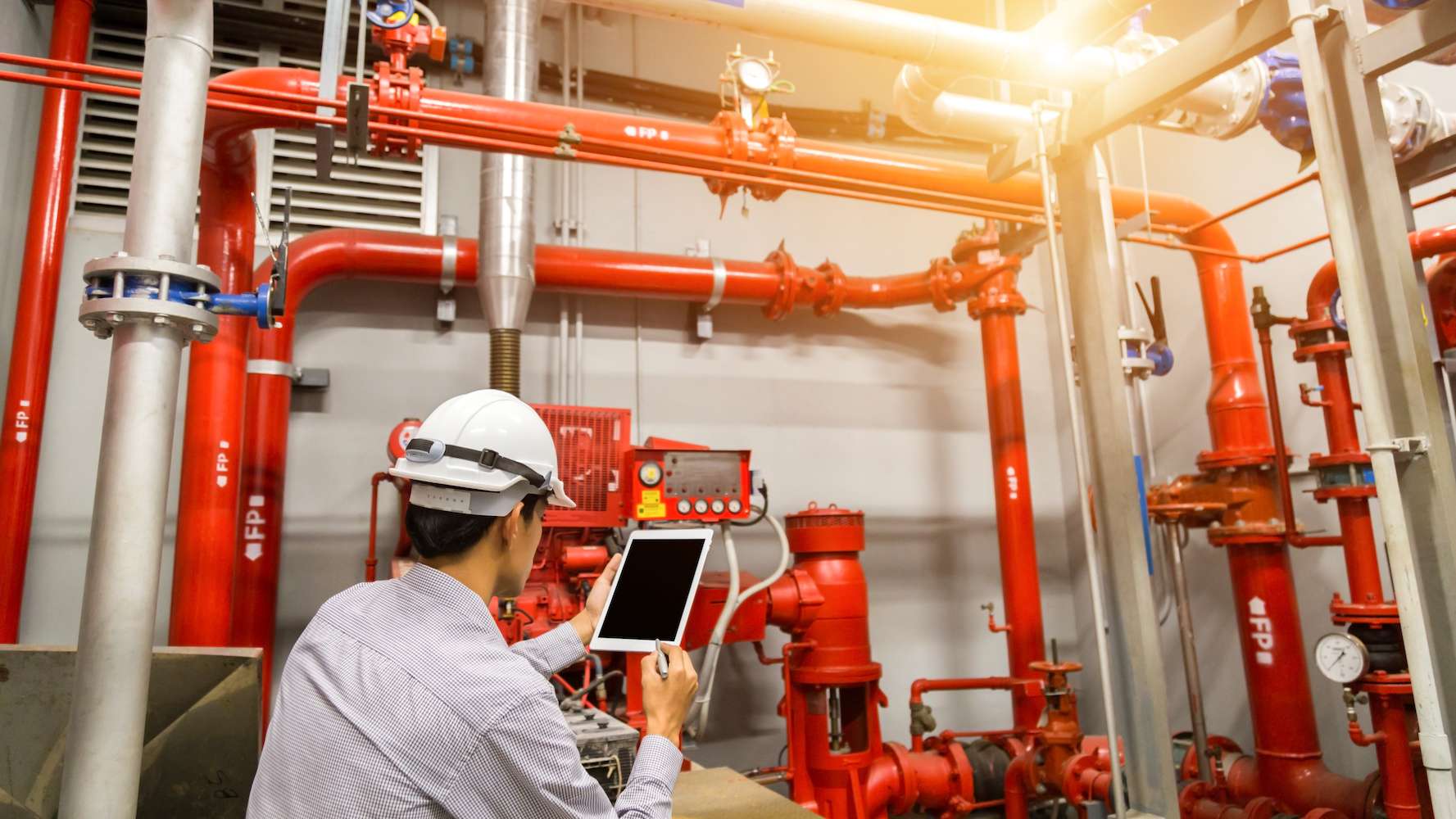 A worker in a hard hat inspects industrial red pipes and machinery, using a tablet for reference in a facility or plant. A worker in a hard hat inspects industrial red pipes and machinery, using a tablet for reference in a facility or plant.