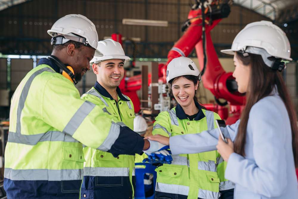Four engineers wearing hard hats and safety gear stand in a factory setting, stacking their hands together in a gesture of teamwork.