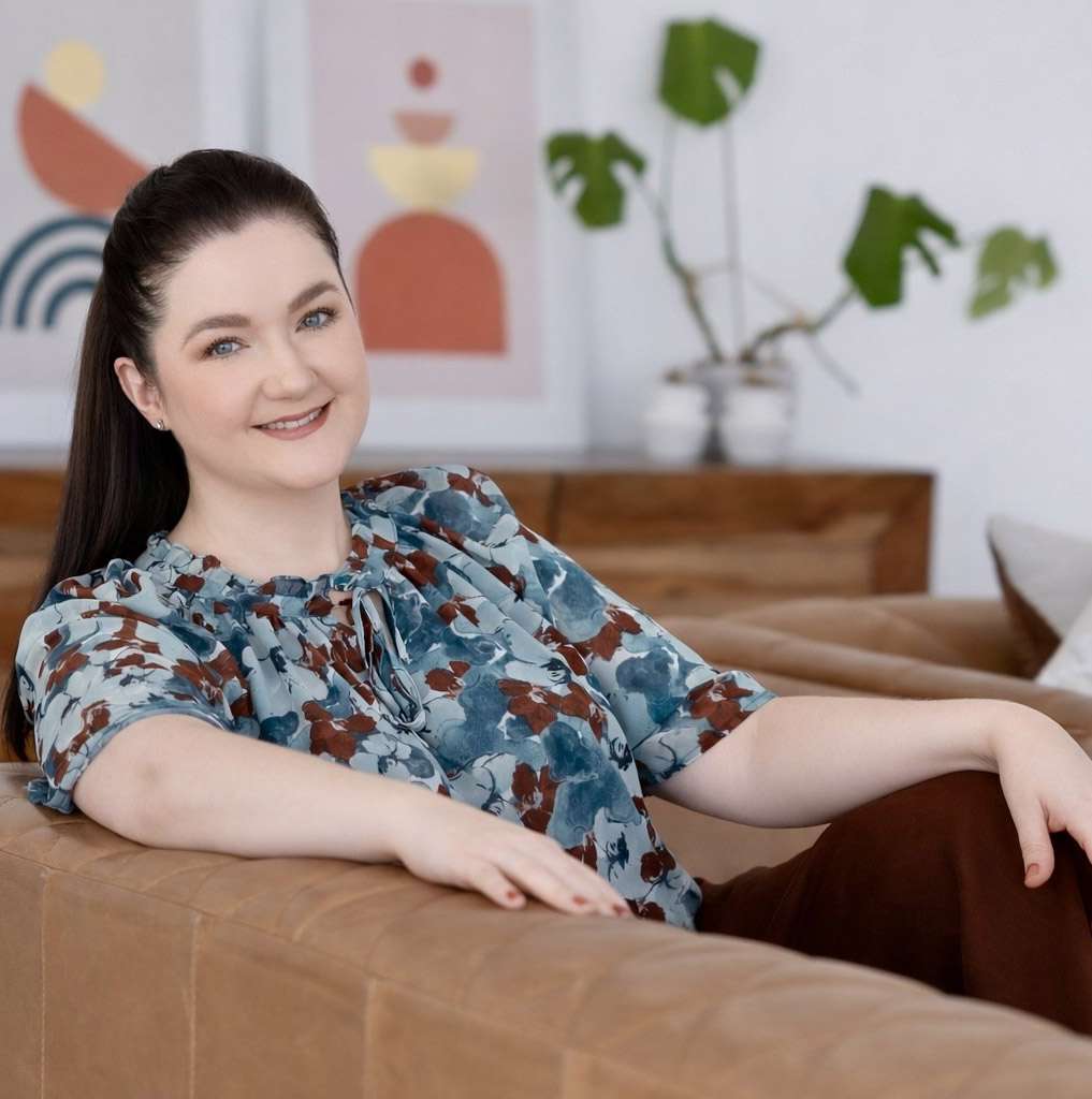 A woman with long dark hair sits on a brown couch, smiling at the camera. Abstract art and a plant are visible in the background. A woman with long dark hair sits on a brown couch, smiling at the camera. Abstract art and a plant are visible in the background.
