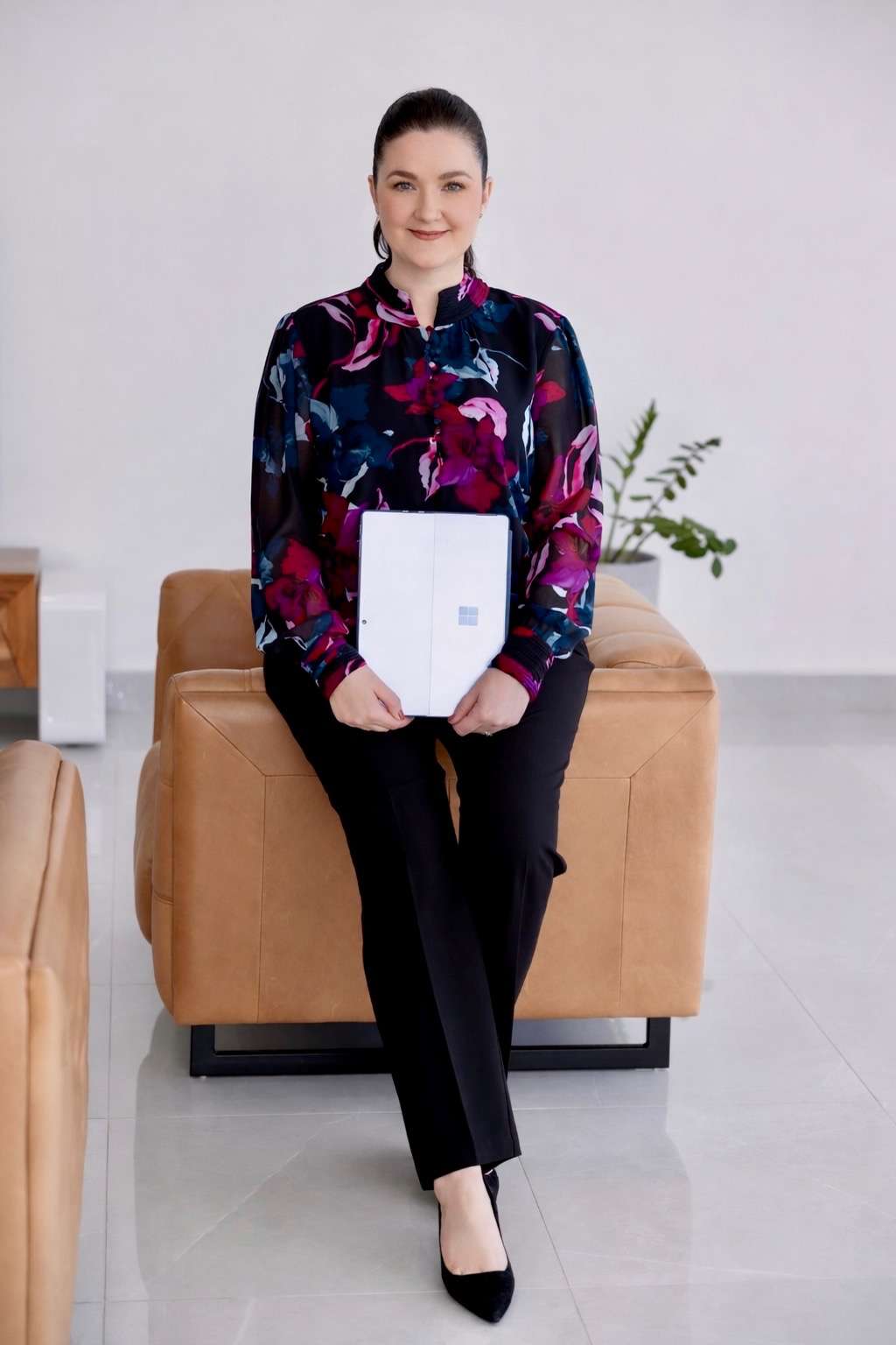 A woman in a floral blouse and black pants sits on a tan ottoman, holding a closed laptop on her lap. A woman in a floral blouse and black pants sits on a tan ottoman, holding a closed laptop on her lap.